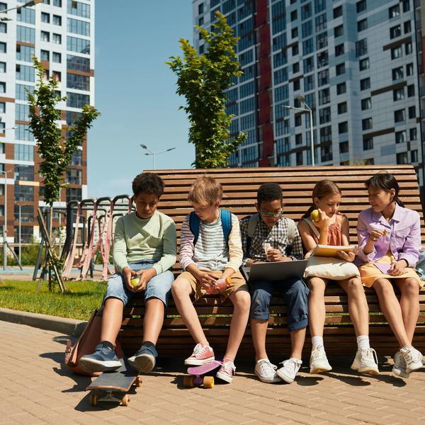 The image shows 5 children sitting on a bench in an urban environment. Some of them are eating, one girl is looking at the screen of her laptop.