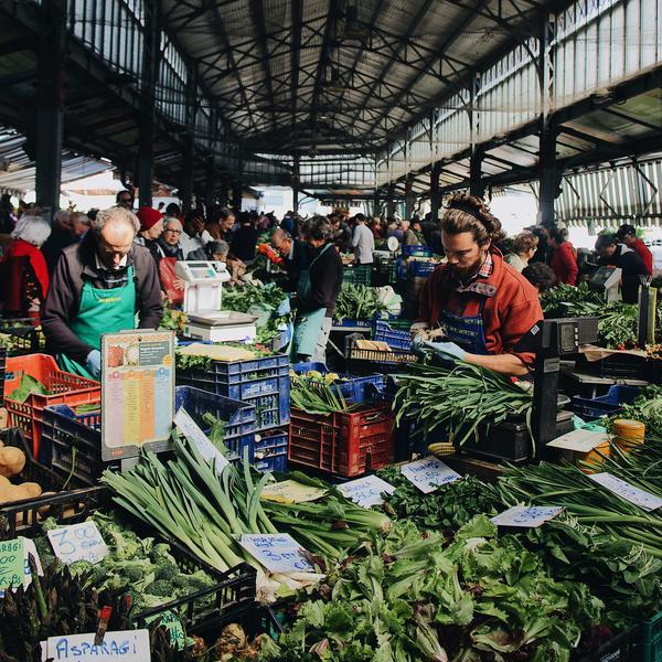 The image shows a food stand with vegetables located in an indoor market hall. Vendors and visitors of the market in the background.